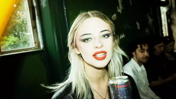 TTSFU wearing green eyeshadow and holding a can. The background features people and is blurred.