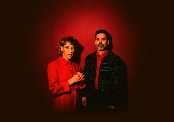 Two members of tune-yards wearing suits on a red background. The image features vibrant colours and dramatic red lighting.