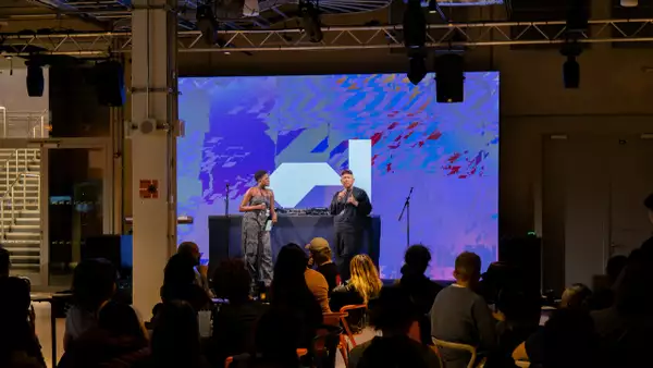 Two people stood on stage in front of DJ decks with the Factory International logo on a purple background on the screen behind them. There are a crowd of people sat on chairs watching them.