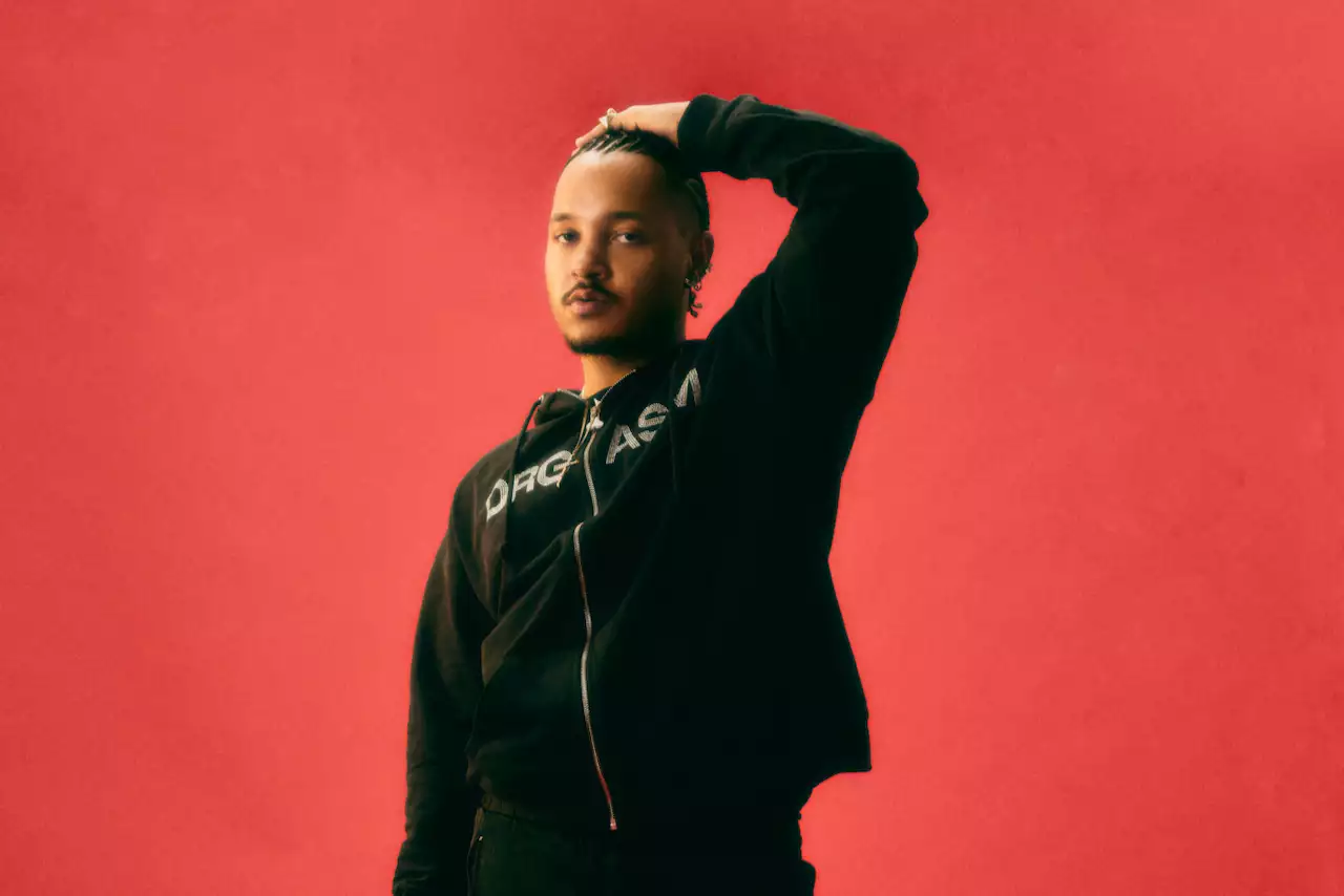 Bennettiscoming wearing a black tracksuit top, on a bright red background. They are holding a hand above their head.