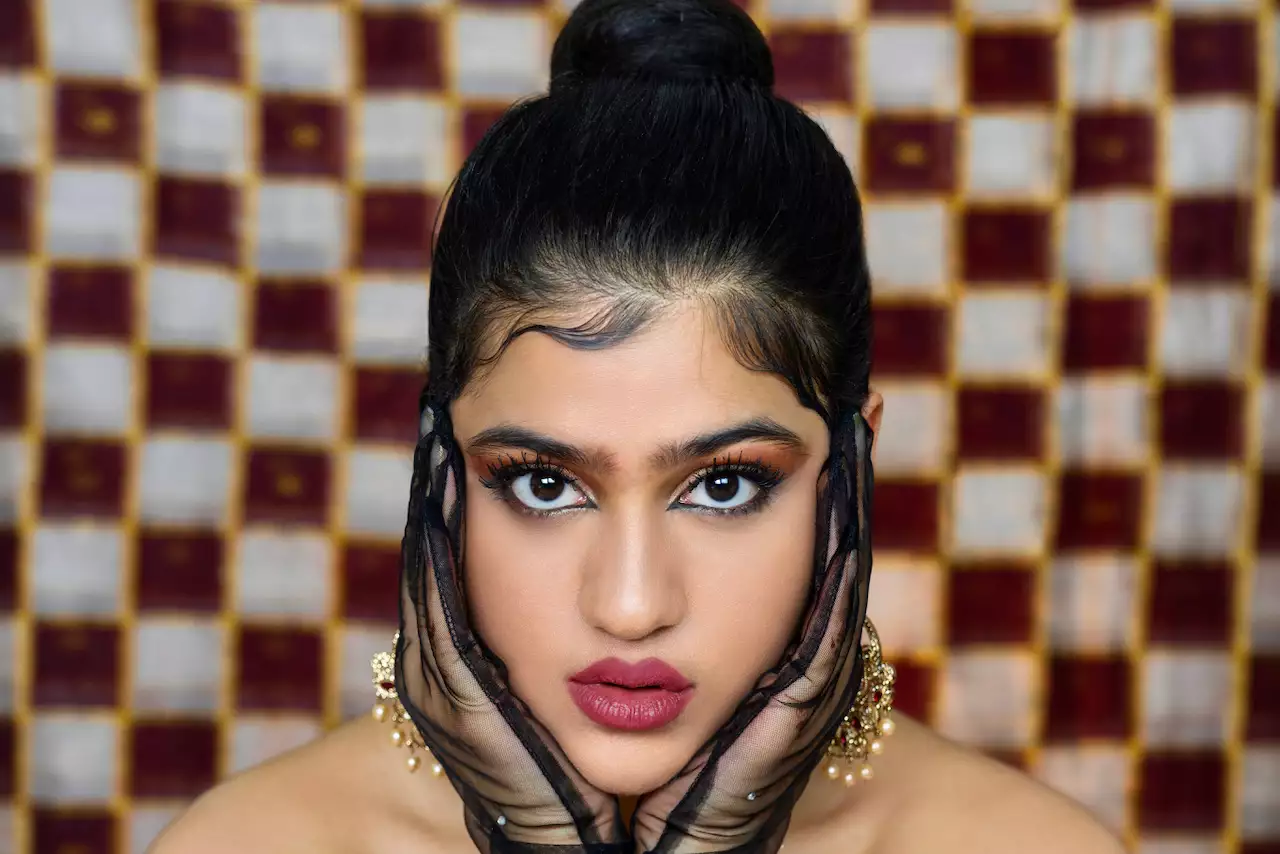 RIYA with their hair in a bun and head in their hands, which are covered with black, opaque gloves. The background is chequered red and white.