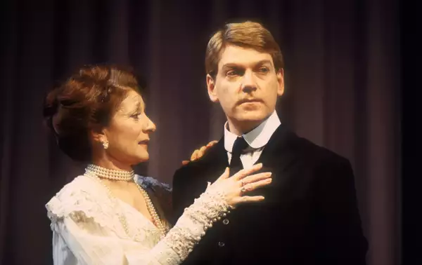 Jane Lapotaire and Kenneth Branagh on stage as Gertrude and Hamlet. Jane is wearing a white lace dress with her hands on Kenneth's chest. He is wearing a suit and looking off-camera.