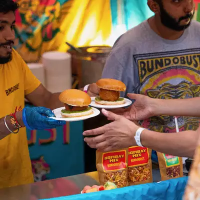 Two people in a Bundobust food truck serving burgers, wearing brightly coloured Bundobust t-shirts