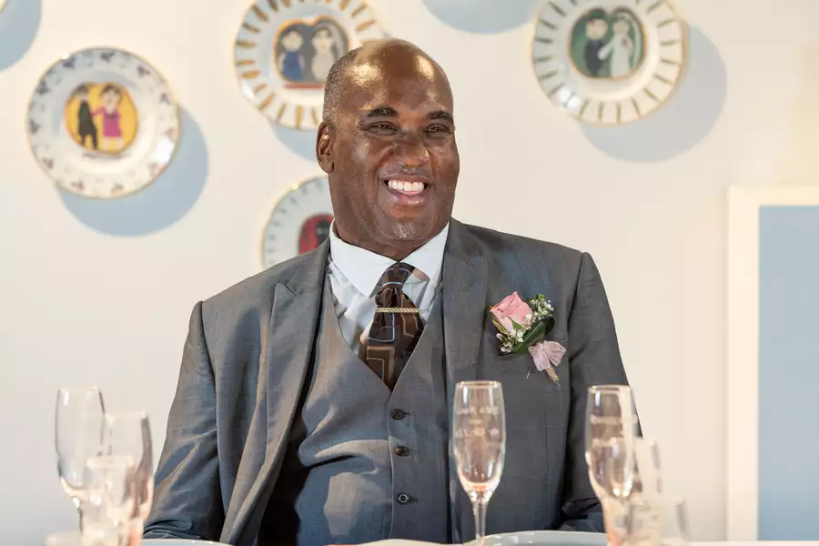 Horace Lindezey in a suit with a pink rose sits at a table with champagne glasses, in front of a wall decorated with illustrated plates.