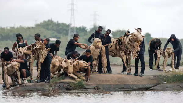 A herd of cardboard animal puppets being operated by people in black clothes by the side of a river