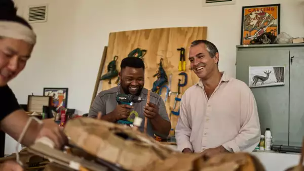 Two people smiling and laughing in a workshop setting for THE HERDS. One is holding a drill.