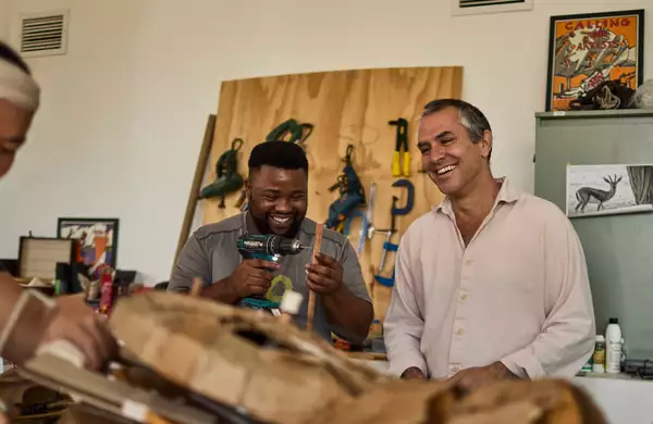 Two people smiling and laughing in a workshop setting for THE HERDS. One is holding a drill.