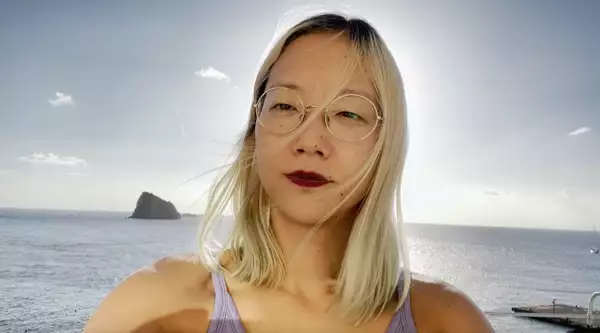 A photograph of Christine Sun Kim, backlit by the sun and standing in front of the sea with a small island above her right shoulder