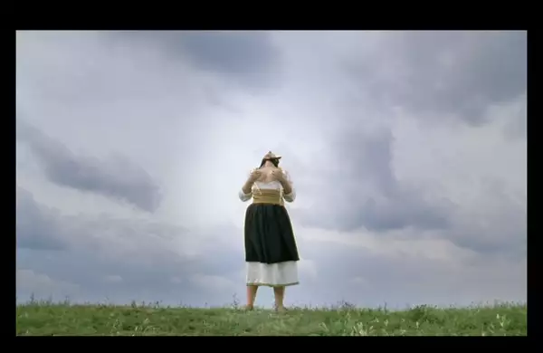 A woman in traditional Eastern European attire stands barefoot in a grassy field under a dramatic, cloudy sky lifting their heads upward in a ritualistic pose