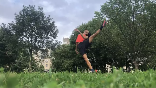 A horizontal photo of ​​Benedict Nguyễn, wearing black shorts, orange tank, and black and orange sneakers, standing on right leg with left leg raised to the side, left arm bent in front of her torso, head tipped back, in a field with green grass and trees, with buildings in distant background.