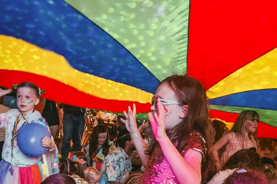 Children stand and play under a large, colorful parachute during an indoor event.