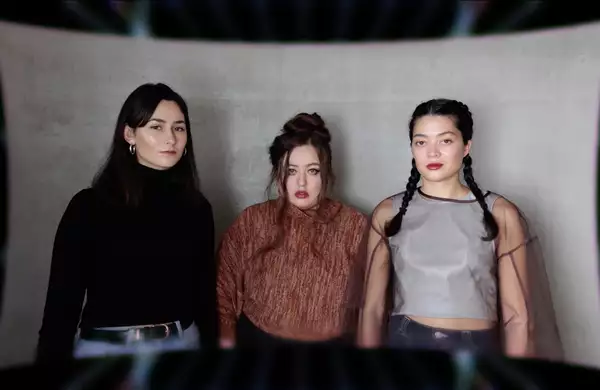 Three members of Keiken staring at the camera, stood in front of a large, grey piece of material.