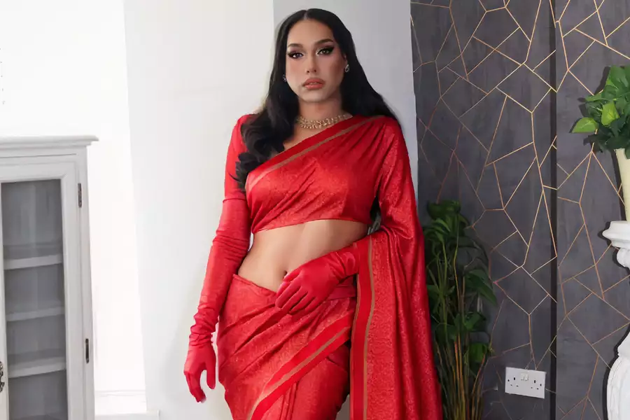 A poised individual stands confidently indoors wearing a striking red saree with gold accents and matching red gloves. Their long, wavy black hair is styled neatly, and they accessorise with gold earrings and a chunky necklace. The backdrop features a geometric-patterned dark wall, a white cabinet, and potted plants, creating a blend of traditional elegance and modern decor.