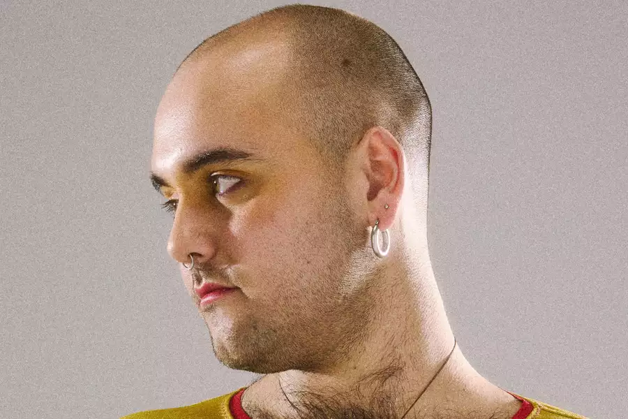 A person with a shaved head gazes to the side against a plain grey background. They wear silver hoop earrings, a nose ring, and a mustard-yellow top with a red collar. Their makeup includes warm yellow eyeshadow around the eyes, and they have subtle facial hair and a chest necklace. The lighting highlights their smooth skin and confident expression.