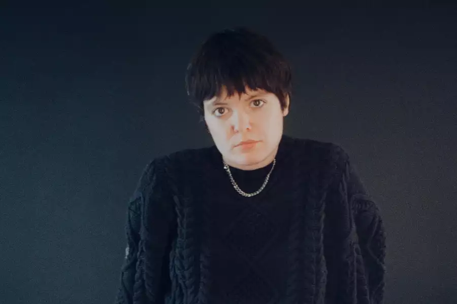 Olivesque sits on the floor in a black jumper against a black backdrop.