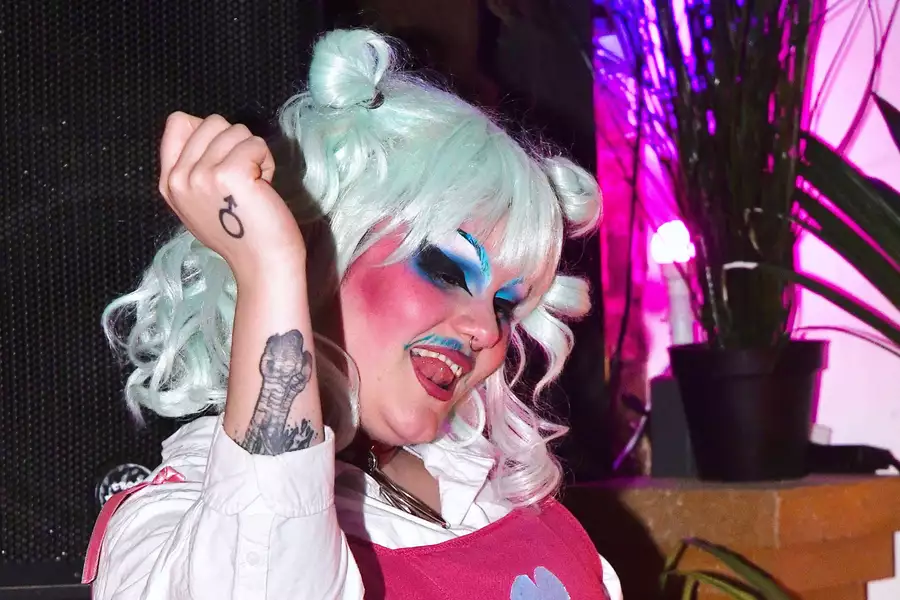 astel mint hair styled in two buns, dramatic blue and pink makeup, and a bold mustache poses playfully with their tongue out and fist raised. They wear a white shirt under a pink dress with a heart on it and display tattoos on their arm. The background features potted plants and vibrant pink-purple lighting.